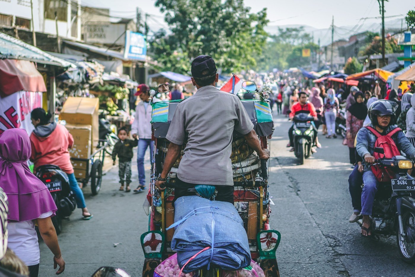A becak driver making his way through a busy market street lined with stalls and motorbikes