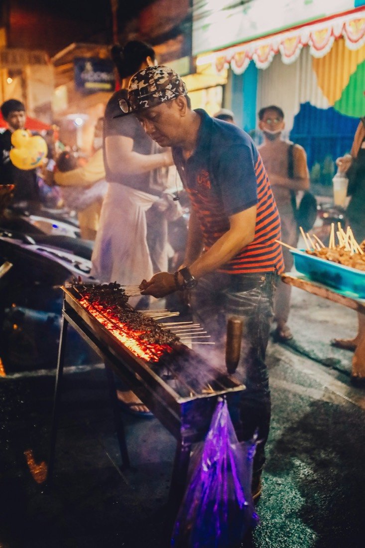 A street vendor grilling satay skewers over coals at a night market